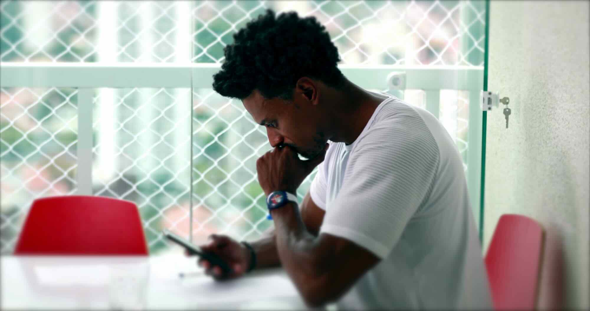 A man sits alone by a window looking at his phone with a thoughtful, weighed-down expression