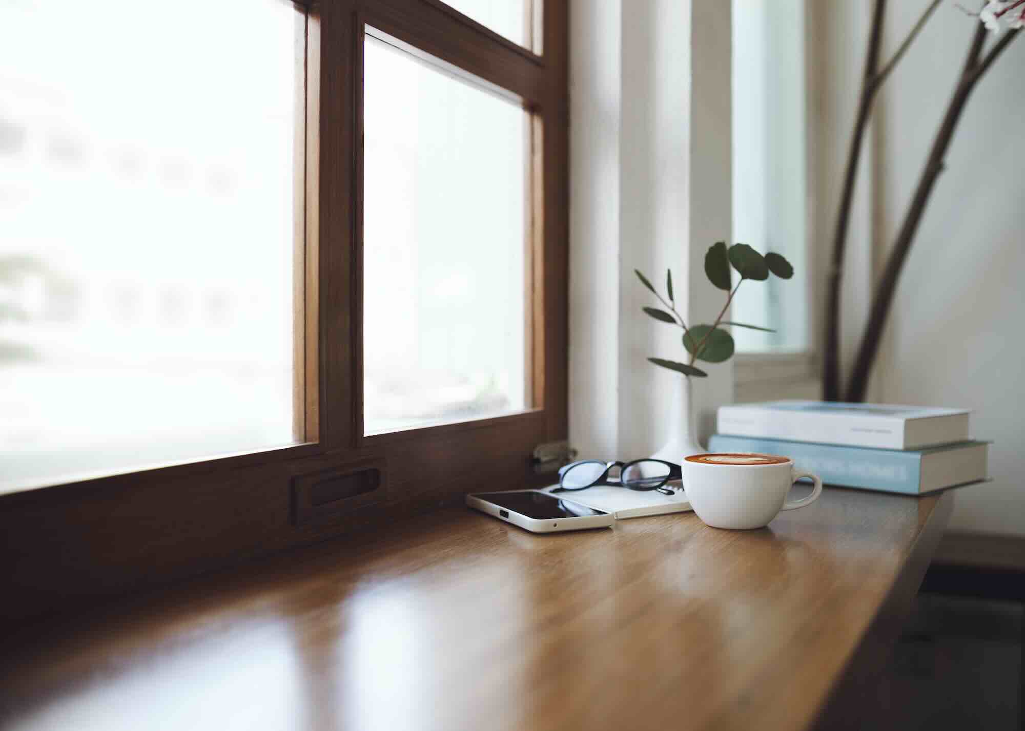 A phone at rest beside reading glasses, a coffee cup, and a small stack of books on a wooden sill in soft daylight