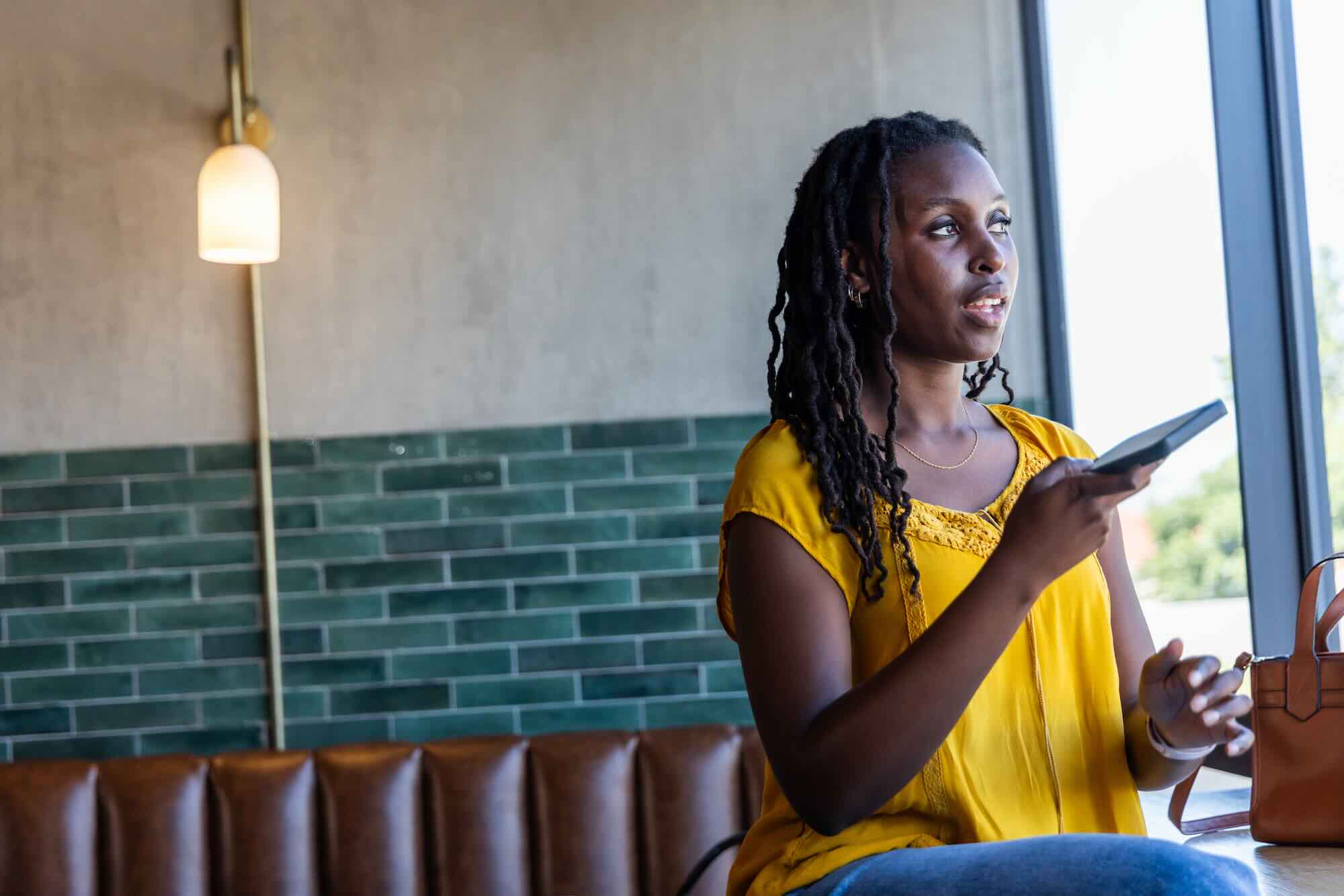 A woman stands by a window holding her phone, looking up with a calm, grounded, decided expression