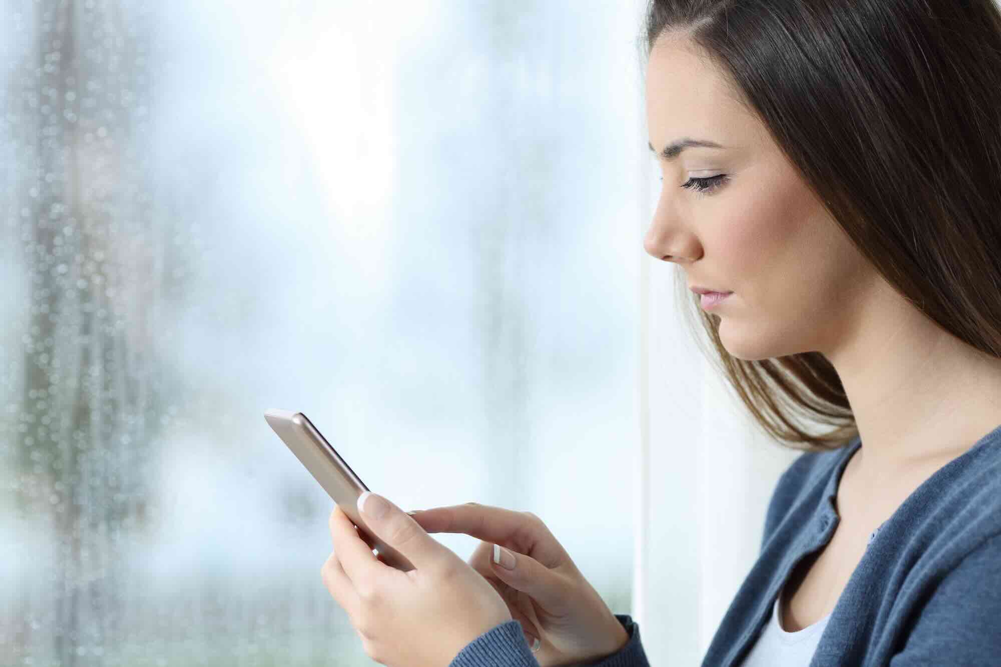 A woman in profile reads a message on her phone with a calm, serious expression beside a rain-streaked window